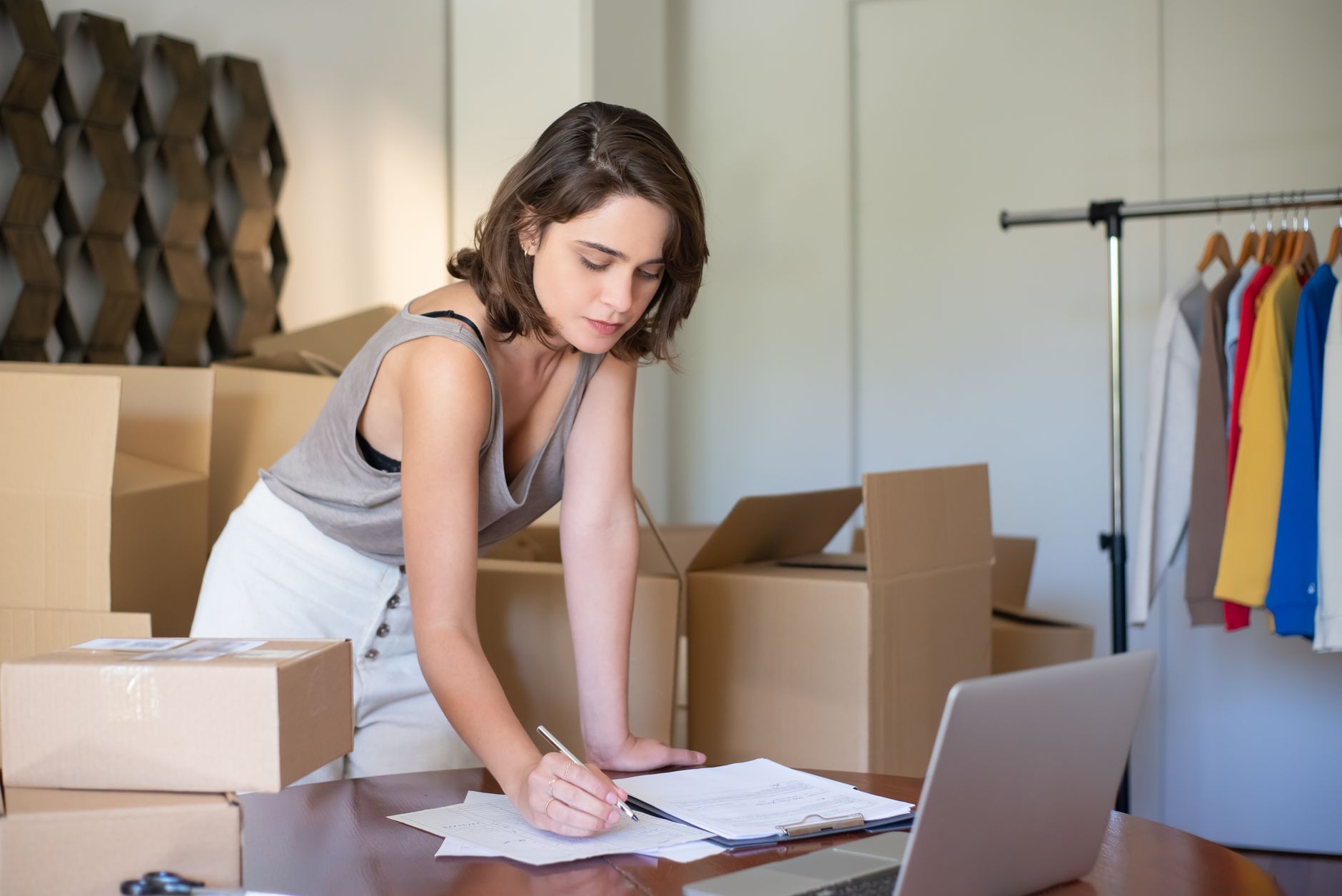 Une jeune femme gère sa boutique de vêtements en ligne depuis son bureau à domicile, entourée de cartons et d'un ordinateur portable