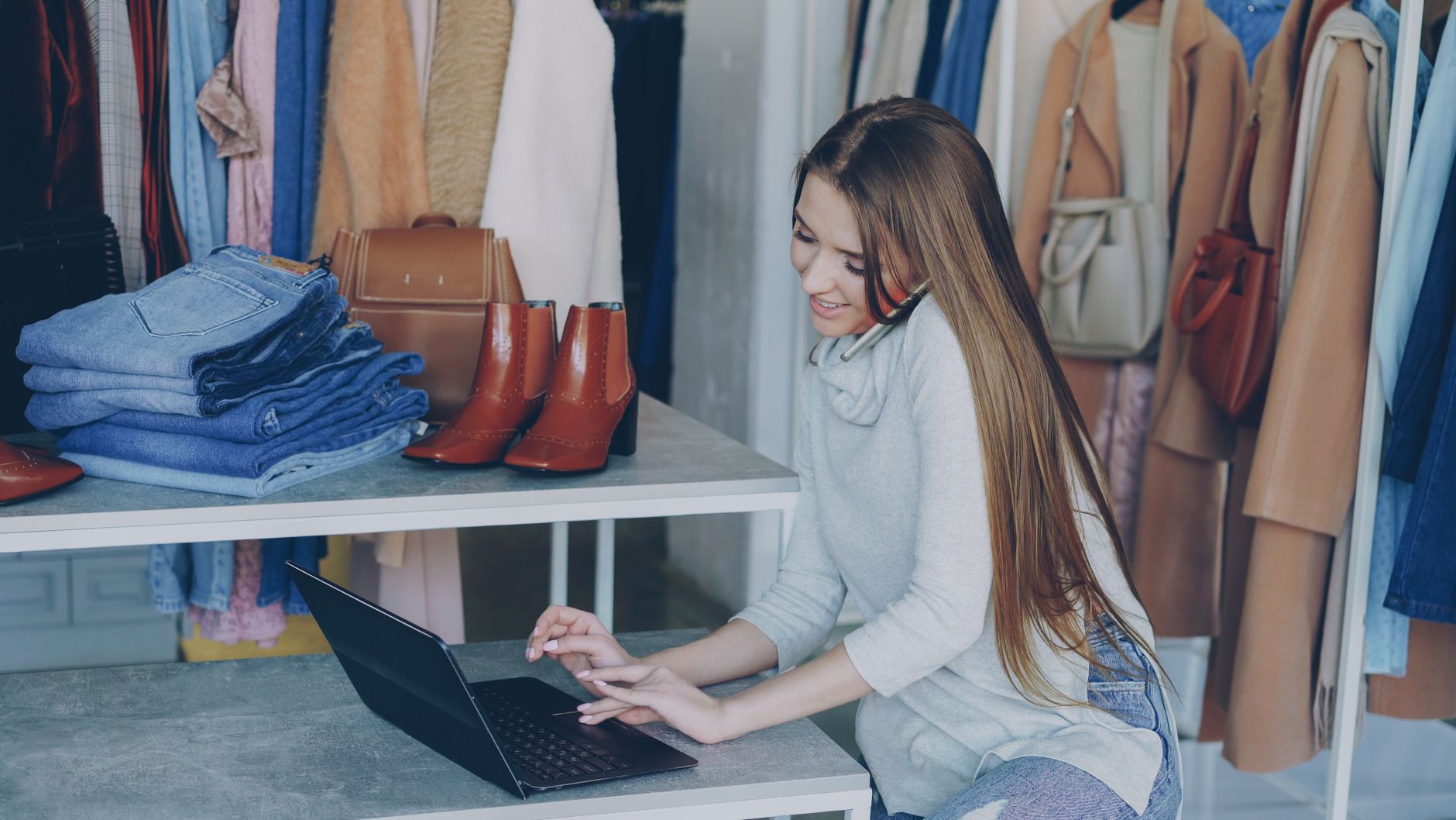 Une commerçante gère sa boutique de vêtements, entourée de pièces et d'un ordinateur portable